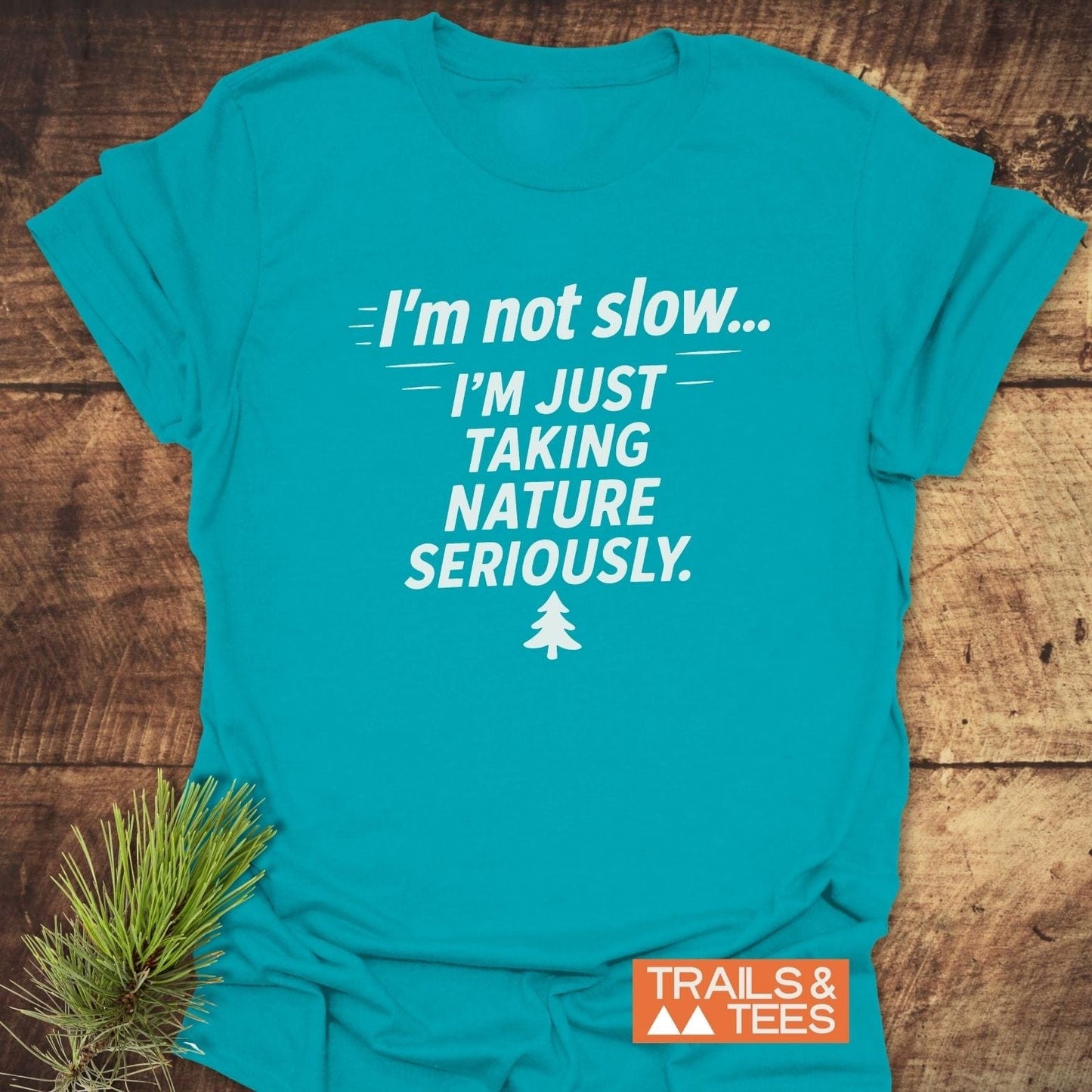 Teal T-shirt with I'm Not Slow... I'm Just Taking Nature Seriously slogan, pine-tree icon, and TRAILS & TEES logo, emphasizing outdoor adventure and comfort.