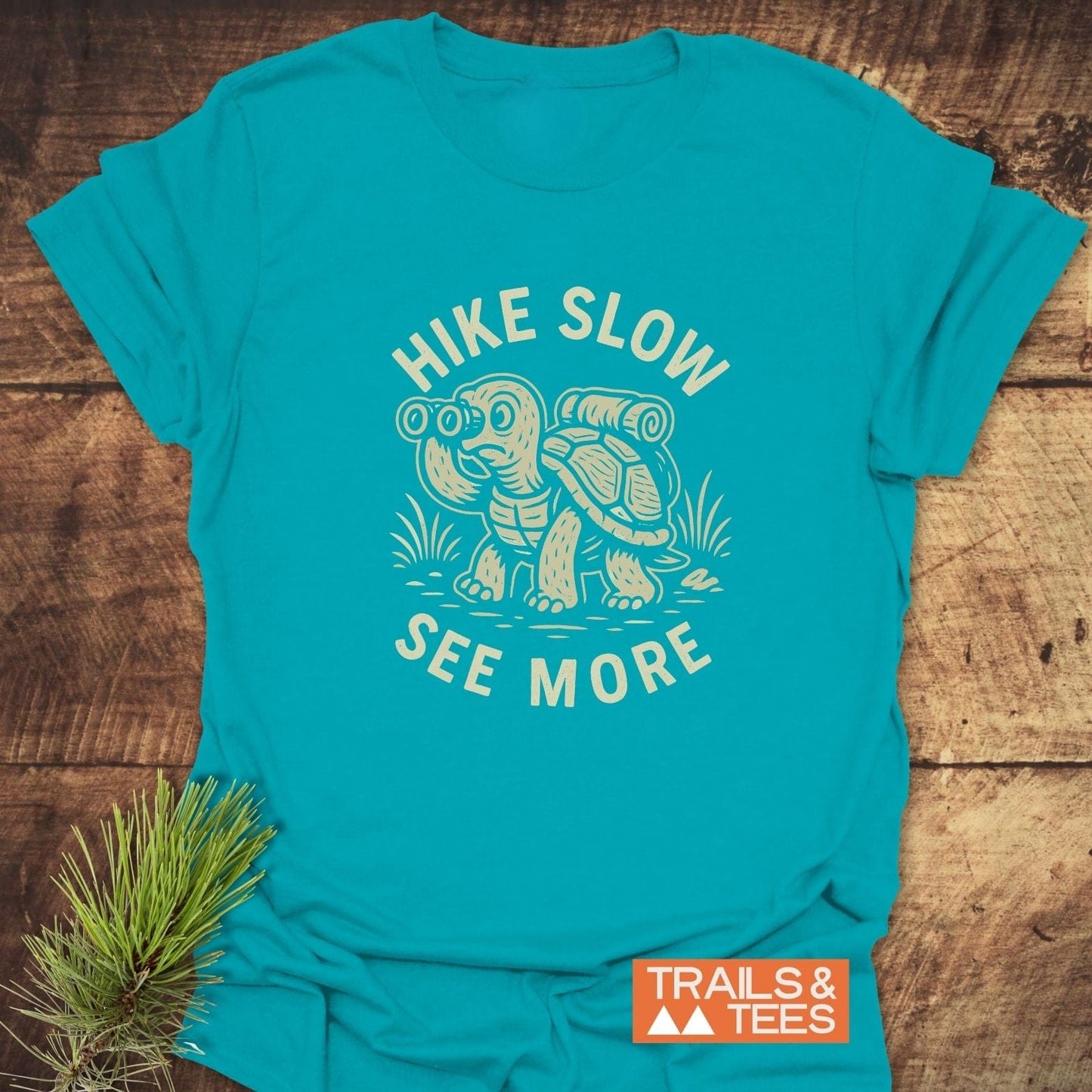 Hike Slow Funny T-Shirt featuring a cartoon tortoise with a backpack and binoculars, slogan “SEE MORE,” and “TRAILS & TEES” logo, on a wooden surface.
