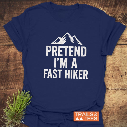 Navy-blue Pretend I'm A Fast Hiker T-shirt with white mountain graphic, laid on wood, featuring an orange TRAILS & TEES logo and pine sprig beside it.