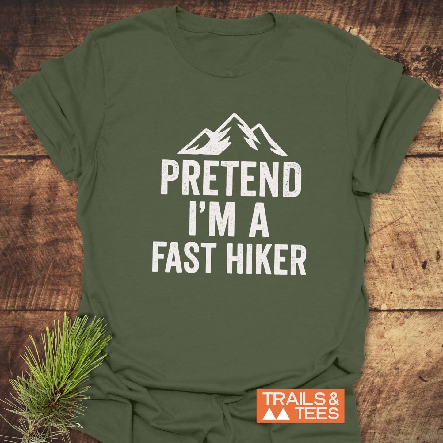 Olive-green 'Pretend I'm A Fast Hiker' T-shirt with mountain graphic, laid on wood. Crafted from soft cotton, pre-shrunk, durable print, by Trails & Tees.