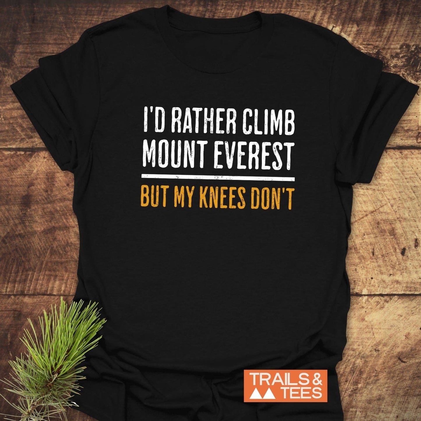 Black T-shirt with I'D RATHER CLIMB MOUNT EVEREST BUT MY KNEES DON'T in bold print. Features Trails & Tees logo, pine sprig accent.