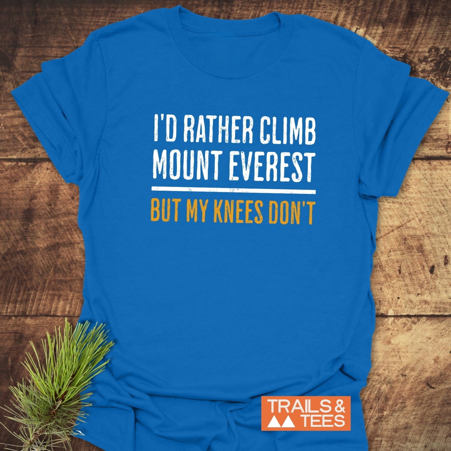 I'd Rather Climb Mt Everest T-Shirt on wood, featuring a white and gold slogan. Includes Trails & Tees logo and pine sprig accent for outdoor vibe.