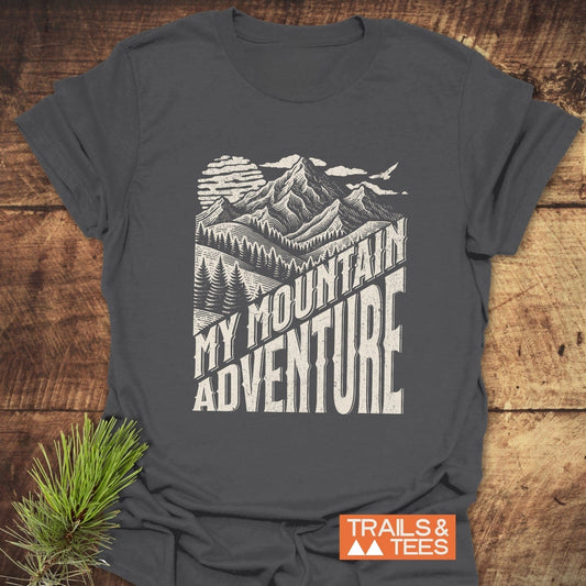 My Mountain Adventure T-Shirt with a mountain graphic and text, crafted from soft cotton for comfort, laid on wood with a pine sprig and logo.