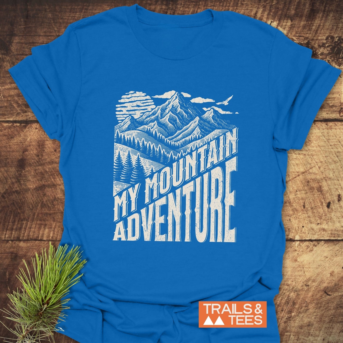My Mountain Adventure T-Shirt features a white mountain design and slogan on blue cotton, laid flat with a Trails & Tees logo.