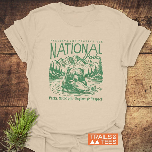 National Parks T-Shirt featuring a bear illustration with forest-green text Preserve and Protect on light-tan fabric, laid beside a pine sprig.