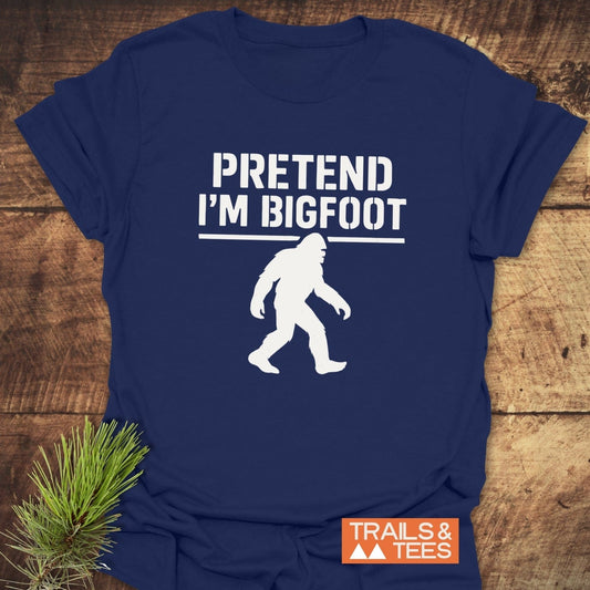 Pretend I'm Bigfoot T-Shirt showcasing a white Bigfoot silhouette and text, laid on wood, featuring durable prints and soft cotton for outdoor comfort.