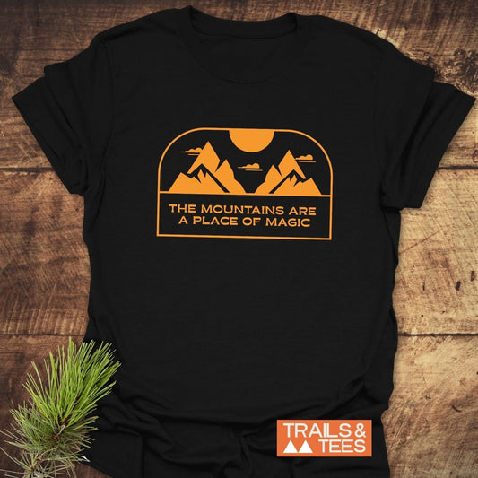 Mountain Magic T-Shirt on wood, featuring bold orange mountain-sun graphic and phrase. Accented by pine sprig, showcasing Trails & Tees logo.