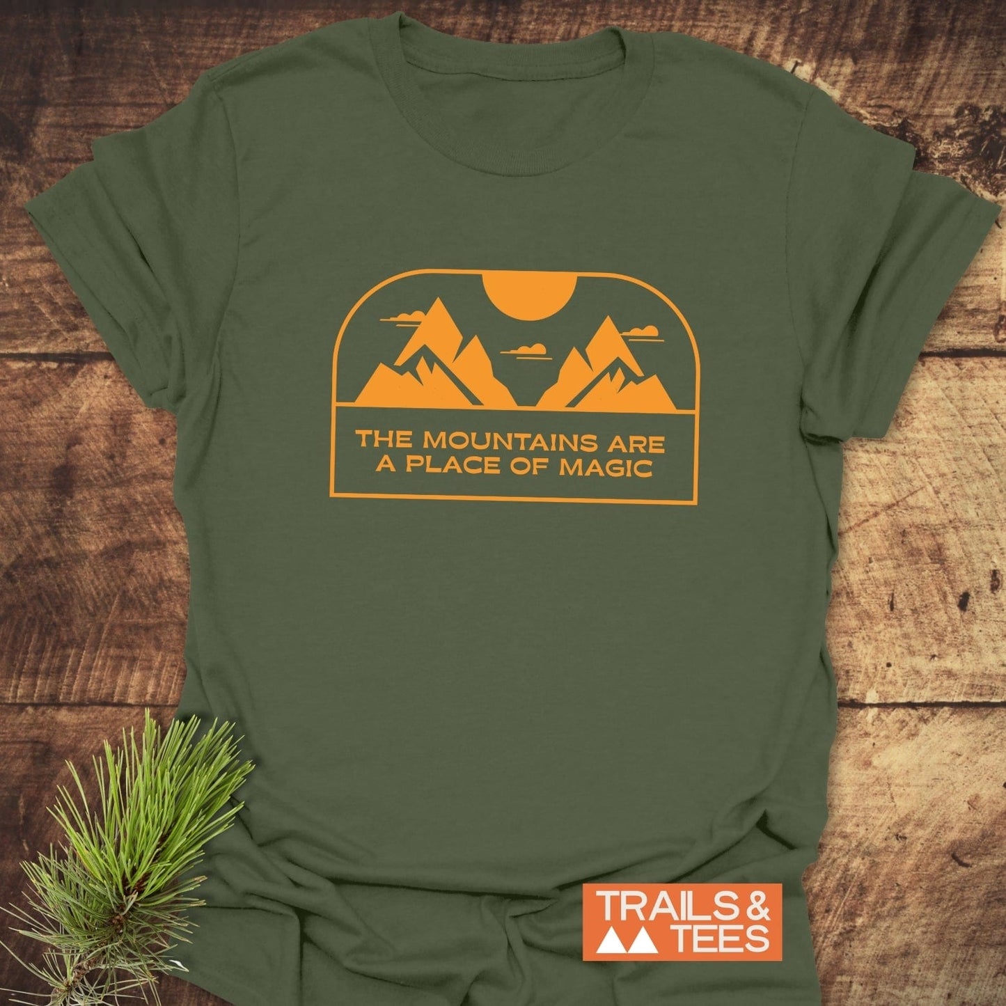 Mountain Magic T-Shirt featuring a stylized mountain print and THE MOUNTAINS ARE A PLACE OF MAGIC, laid on wood with a pine sprig.