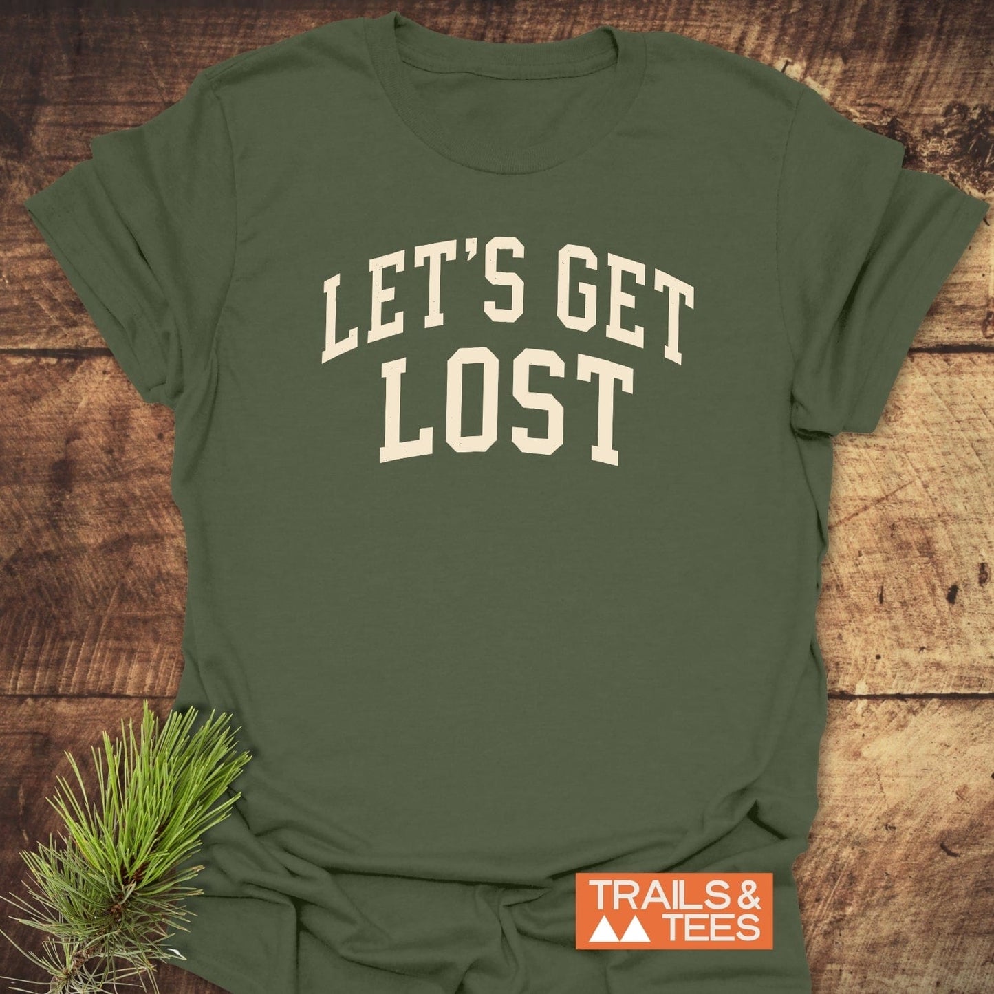 Let's Get Lost T-Shirt on wooden surface, featuring a bold slogan. Includes an orange TRAILS & TEES label and a pine branch accent.
