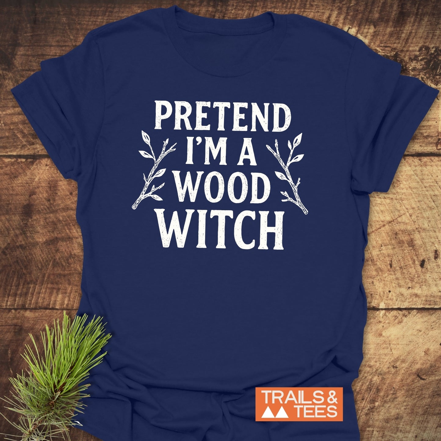 Wood Witch Funny T-Shirt with PRETEND I'M A WOOD WITCH print, laid on wood, adorned with twig illustrations and TRAILS & TEES logo beside a pine sprig.