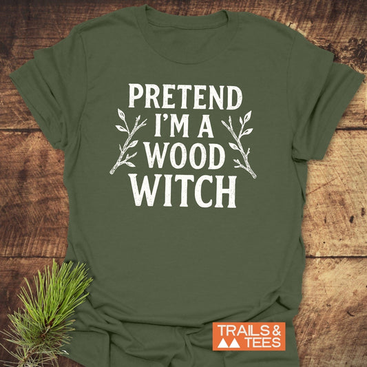 Wood Witch Funny T-Shirt with “PRETEND I’M A WOOD WITCH” text, twig illustrations, and Trails & Tees logo on a wooden surface with pine sprig.