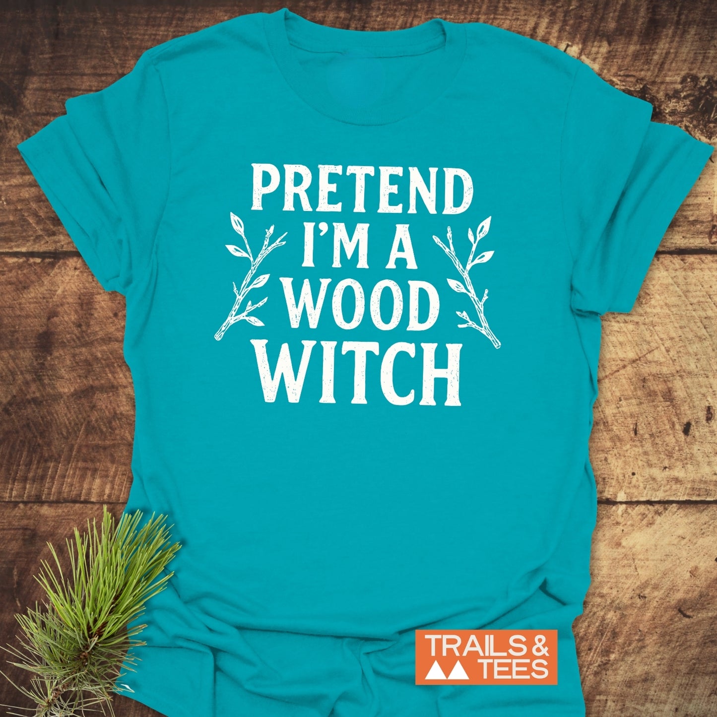 Wood Witch Funny T-Shirt with bold white lettering and twig illustrations, laid on wood surface. Features durable print, soft cotton, and adventure-ready design.