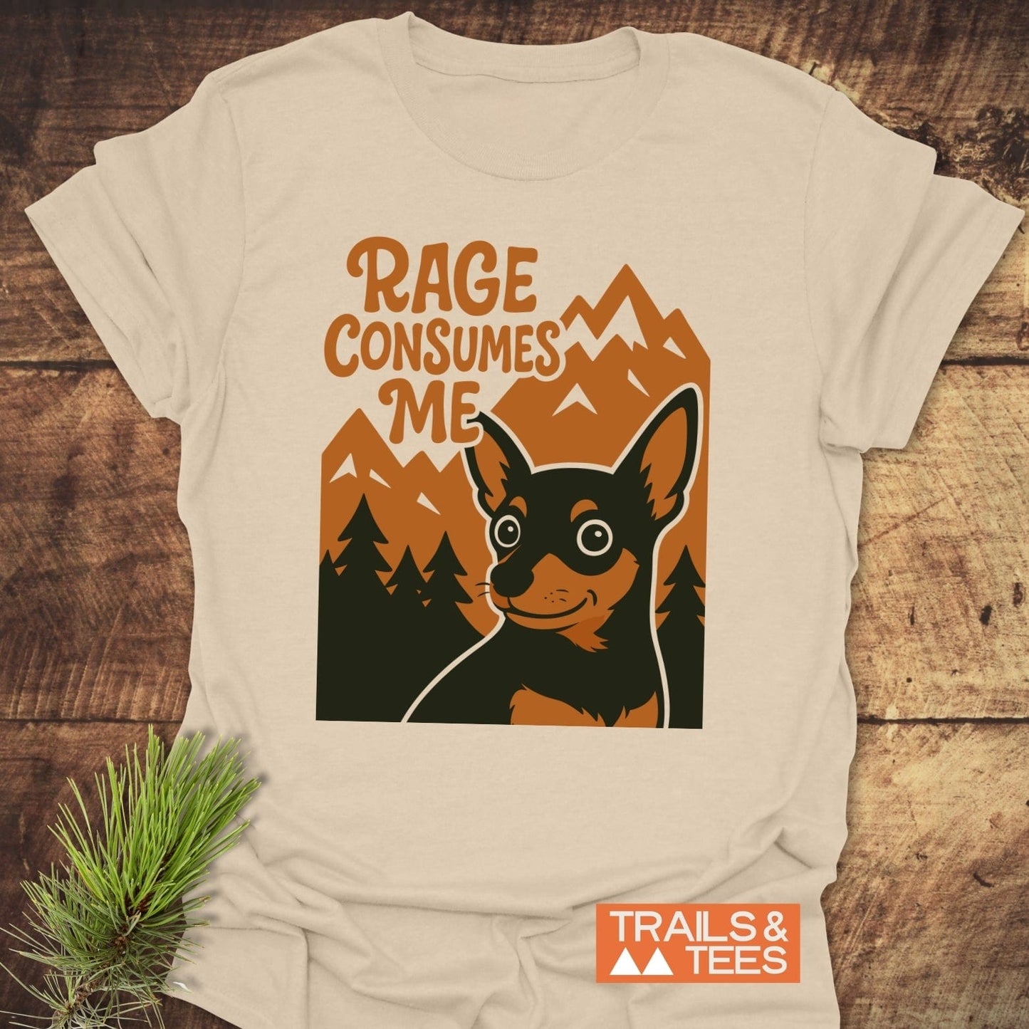 Rage Funny Dog T-Shirt featuring a graphic of a wide-eyed dog amid pine trees and mountains, laid flat on a wooden surface.