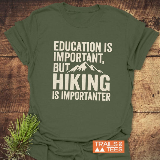 Olive-green Education Is Important T-Shirt with mountain silhouette and hiking slogan, made from soft cotton, laid on wood surface with pine branch beside it.