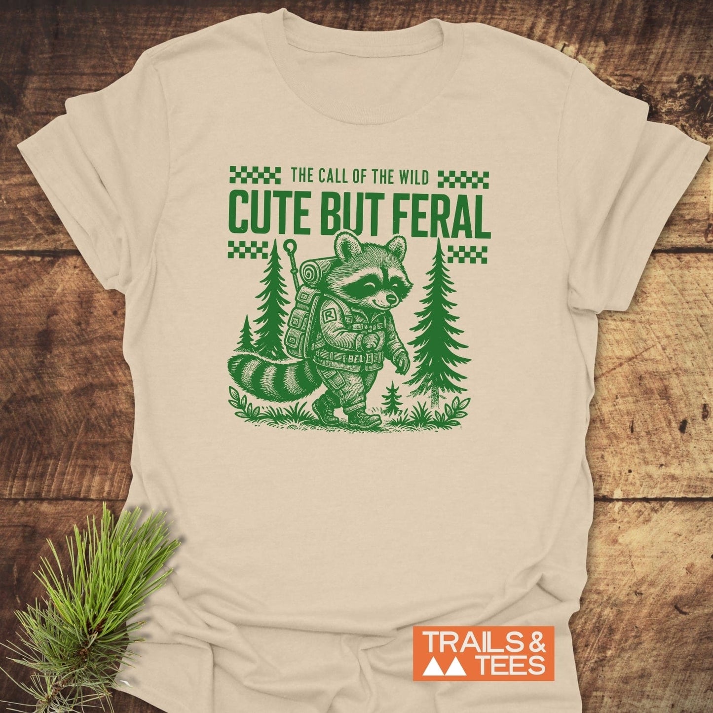 Cute But Feral T-Shirt featuring a cartoon raccoon with a backpack, highlighting trail-ready comfort and durable prints, perfect for outdoor enthusiasts.
