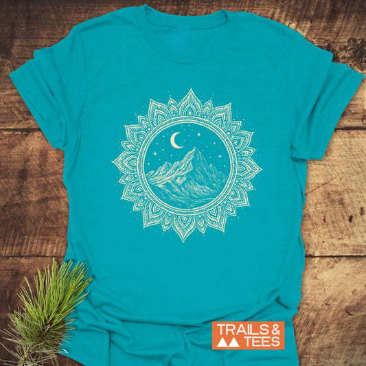 Mountain Mandala T-Shirt featuring a white mandala design with mountains and stars, laid on wood with pine sprig, showcasing outdoor-inspired style.