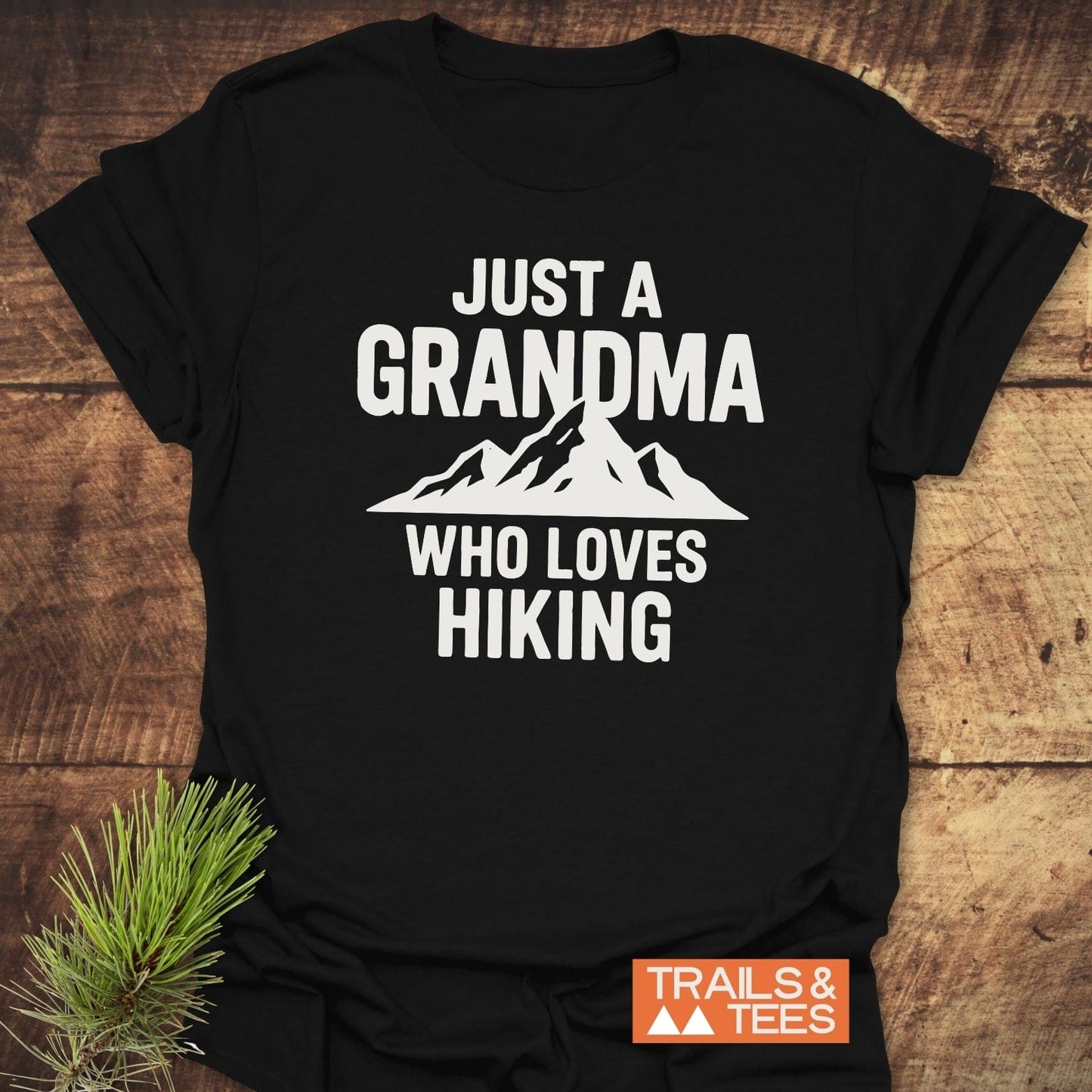 Black T-shirt with Just a Grandma Who Loves Hiking in white, featuring a mountain graphic and Trails & Tees label on a wooden surface.