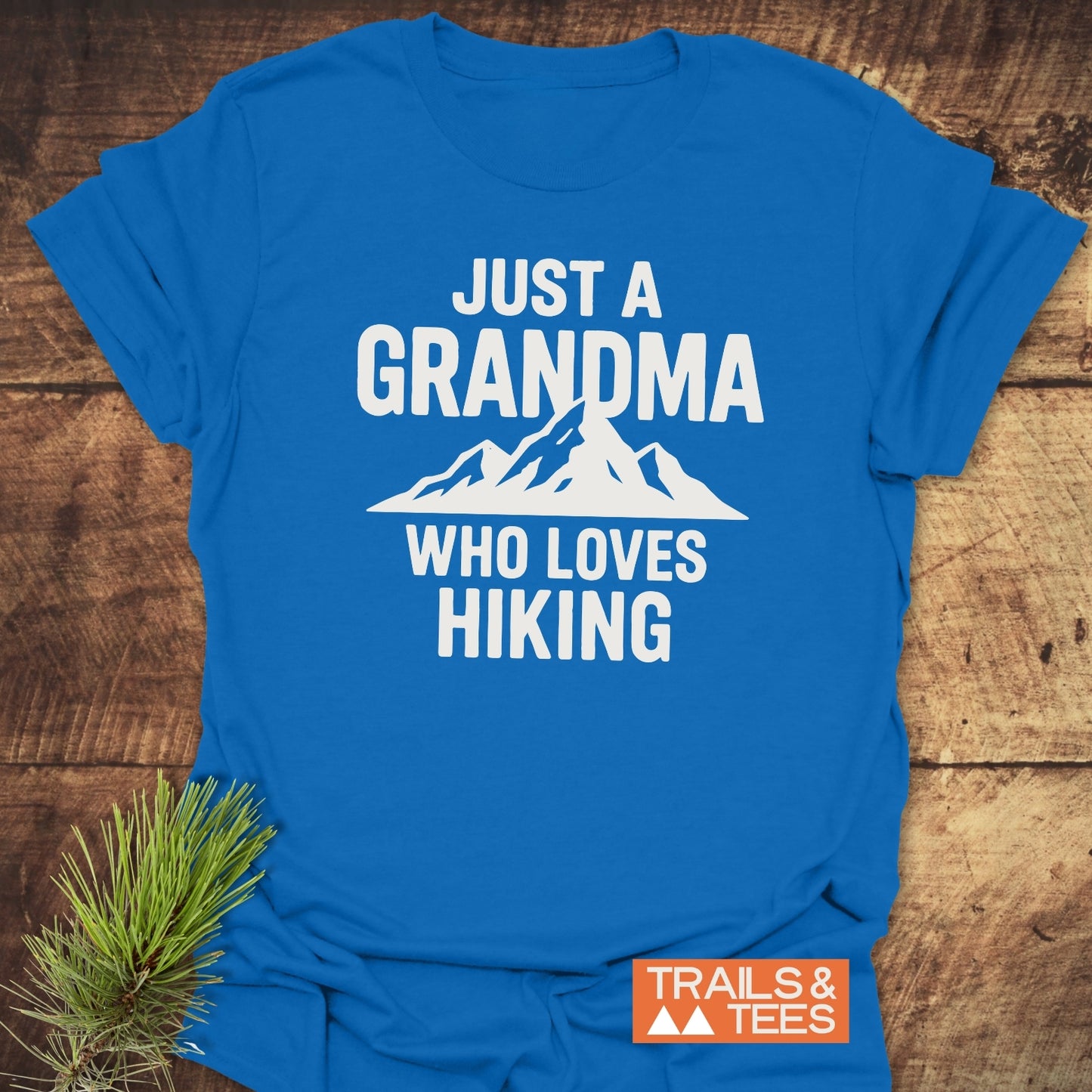 Grandma Loves Hiking T-Shirt featuring a mountain graphic and text, with a Trails & Tees logo, displayed on a wooden surface with a pine sprig.