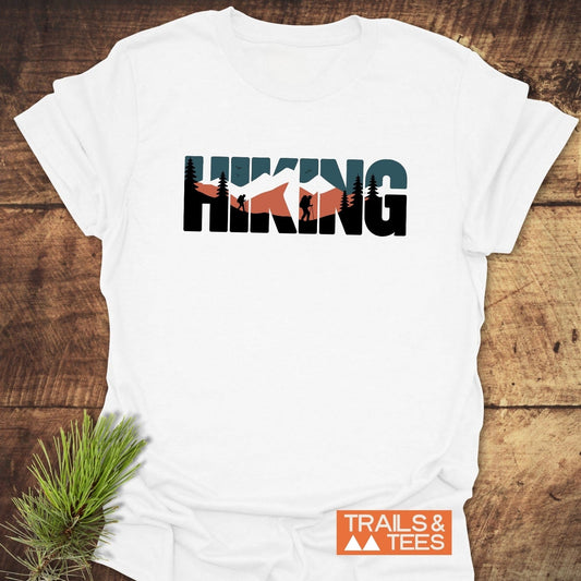 Hiking Retro T-Shirt featuring mountain and forest scenes on letters, laid on wood. Showcases “TRAILS & TEES” logo, crafted for comfort and adventure.