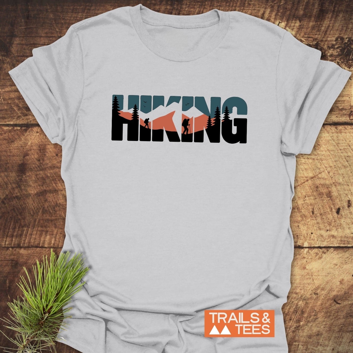 Hiking Retro T-Shirt with mountain and forest scene on chest, showcasing HIKING in muted tones. Includes Trails & Tees logo and pine sprig accent.