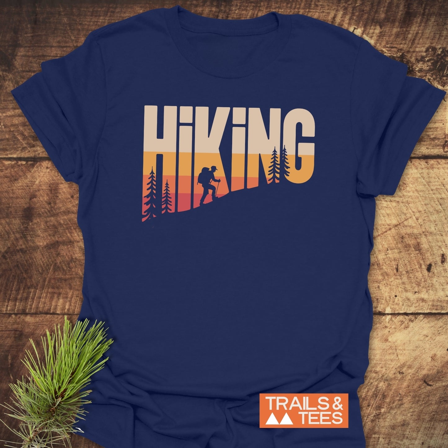 Navy-blue Hiking Vintage T-Shirt with sunset gradient HIKING print, featuring pine trees and backpacker silhouettes. Includes Trails & Tees logo and pine needles accent.