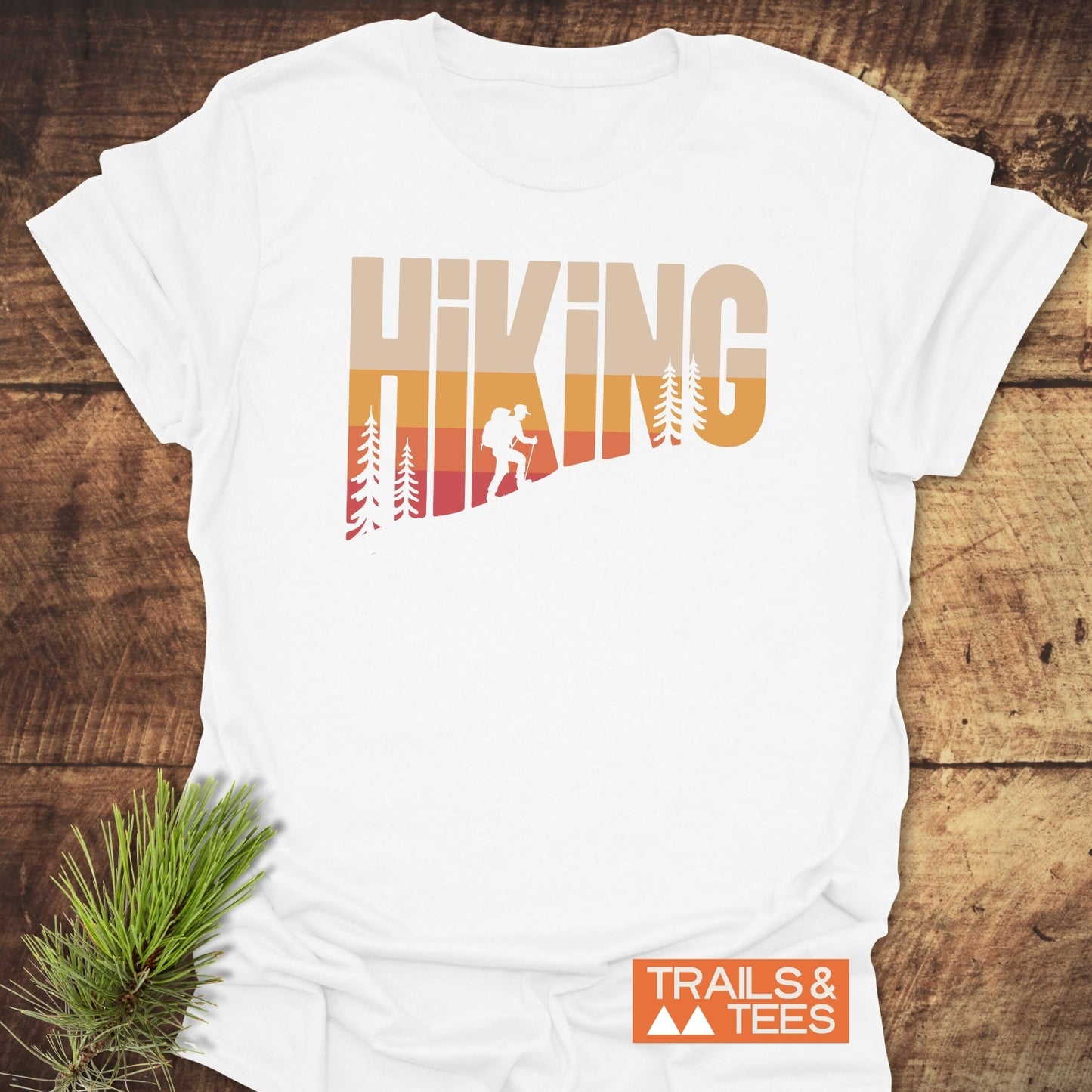 Hiking Vintage T-Shirt featuring a sunset-style “HIKING” print with pine trees and backpacker silhouette, laid on a wooden surface beside a pine sprig.