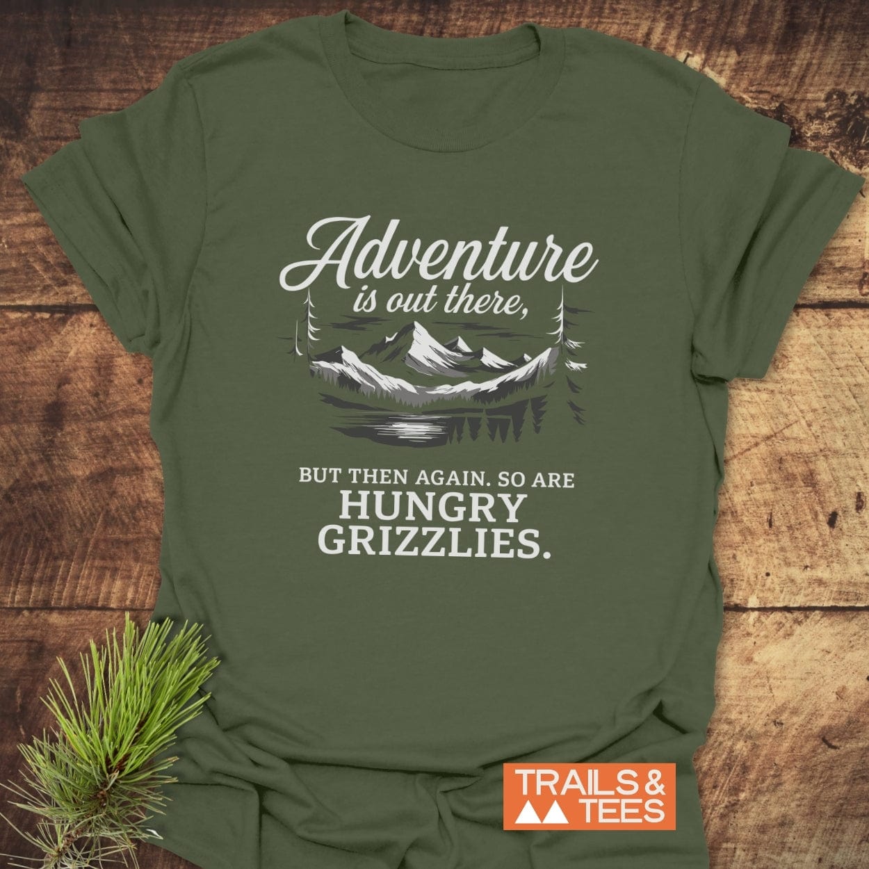 Forest-green T-shirt featuring Adventure Is Out There mountain graphic, with a humorous grizzly warning. Made from soft, durable cotton by TRAILS & TEES.