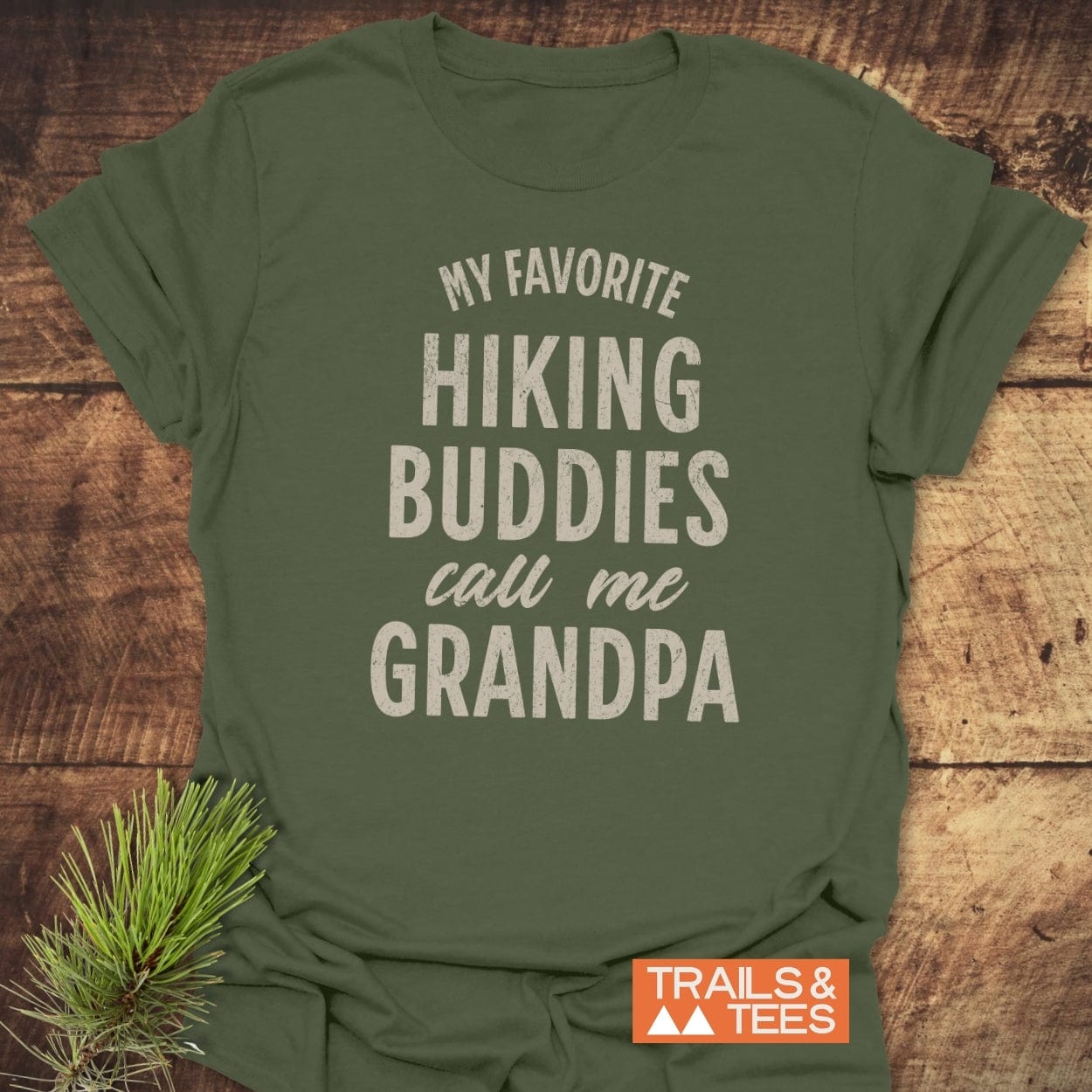 Hiking Grandpa T-Shirt with slogan in distressed white font, folded on wood, featuring Trails & Tees logo, pine sprig beside it.
