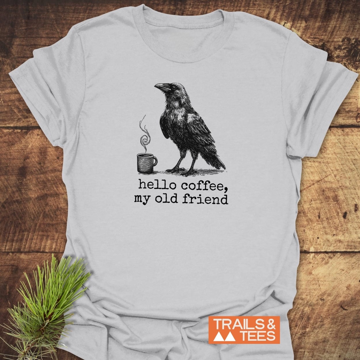 Hello Coffee Funny T-Shirt featuring a crow and coffee mug illustration with the caption, “hello coffee, my old friend,” branded by TRAILS & TEES.