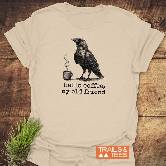 Cream T-shirt with crow and coffee mug design, text “hello coffee, my old friend.” Features durable print and trail-tested comfort from Trails & Tees.
