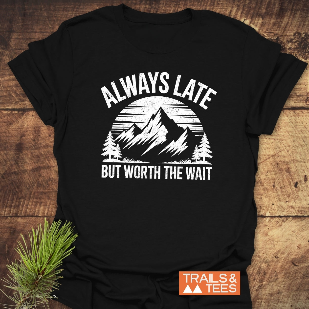 Always Late Funny T-Shirt featuring a mountain scene and slogan, highlighting comfort and durability, set against rustic wood with a pine sprig accent.