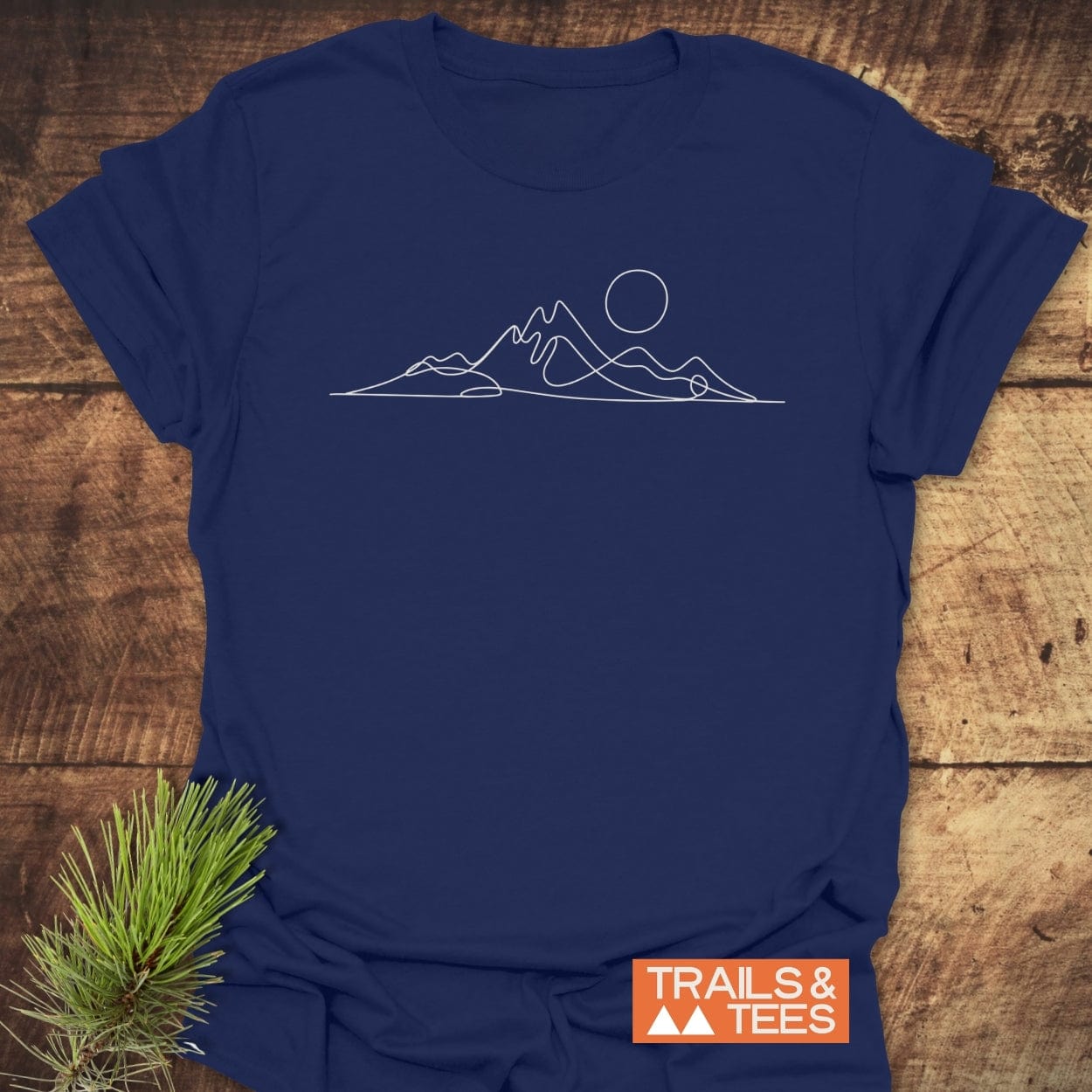Minimalist Mountains T-Shirt with a white mountain and sun graphic on navy cotton, laid on wood, showcasing durable, adventure-ready design by TRAILS & TEES.