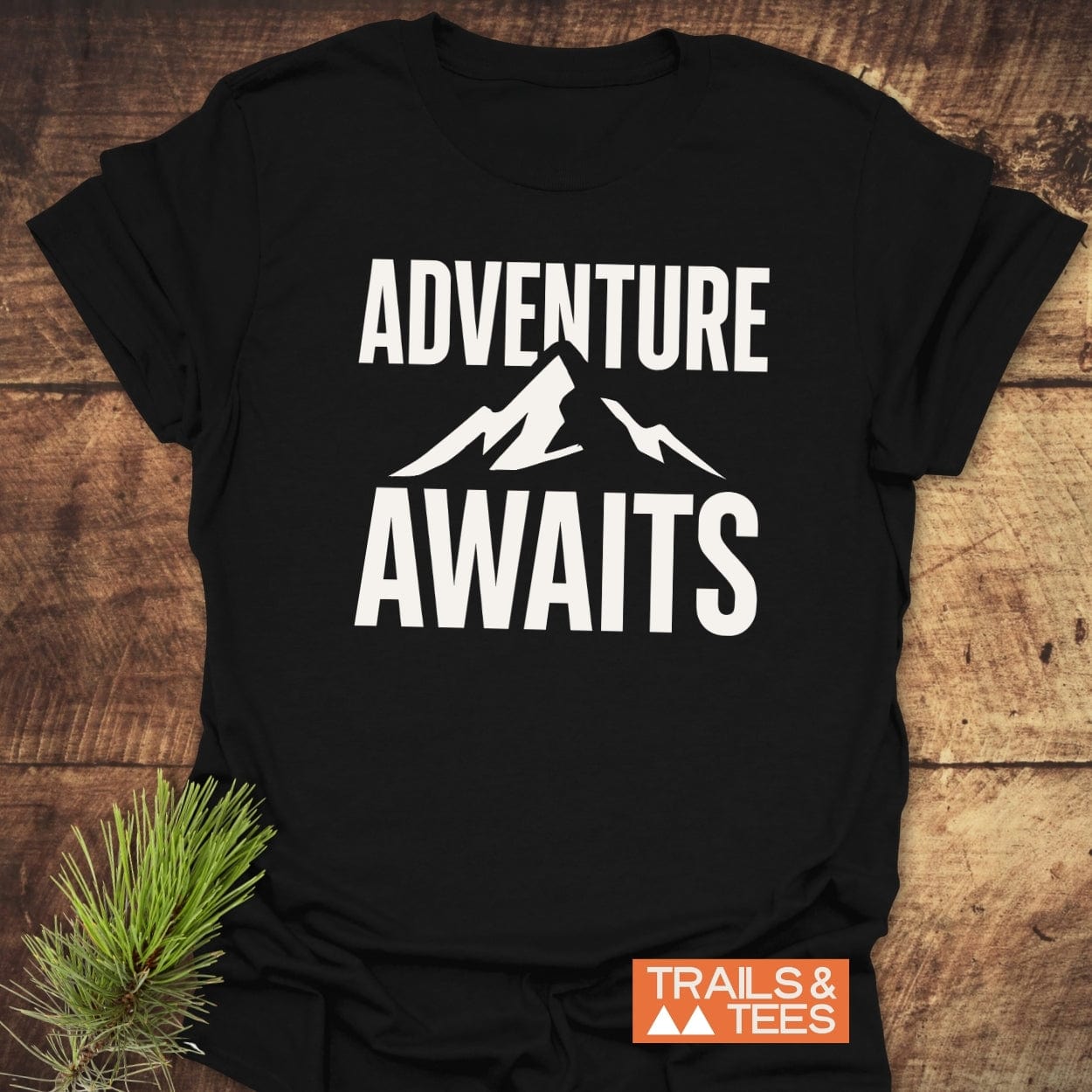 Adventure Awaits T-Shirt on wood, featuring bold ADVENTURE AWAITS text and mountain graphic, with TRAILS & TEES logo, highlighting outdoor-inspired comfort and durability.