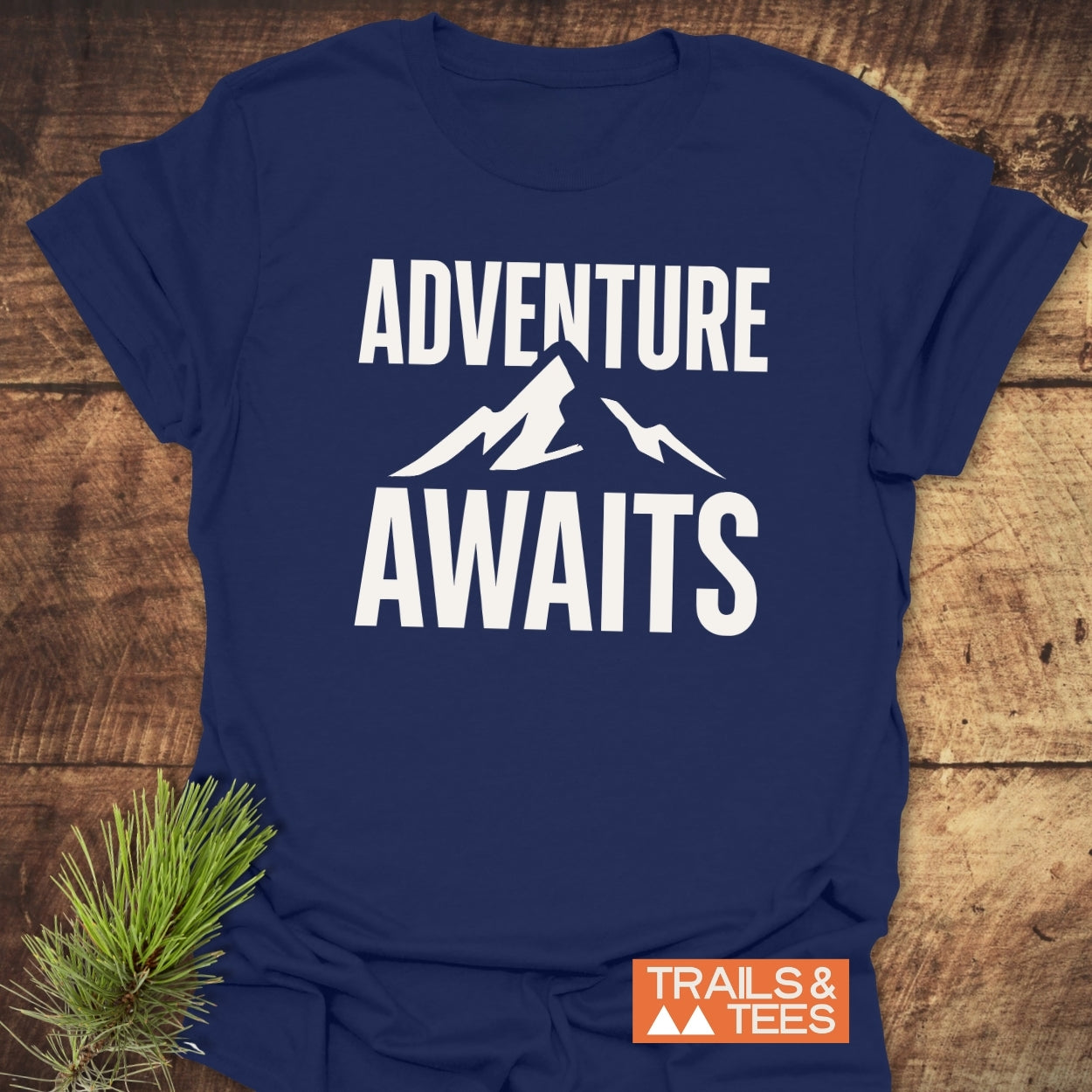 Adventure Awaits T-Shirt featuring bold slogan and mountain silhouette, crafted from soft cotton with durable prints. Includes Trails & Tees logo and pine sprig accent.