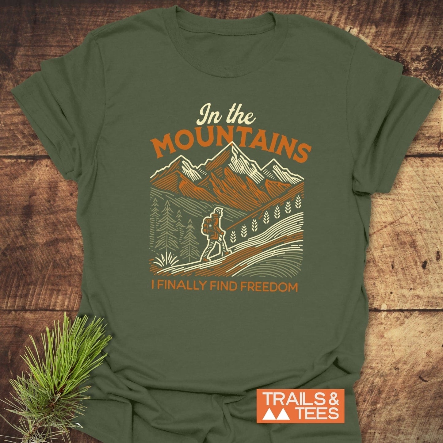Mountain Freedom T-Shirt with mountain scene and lone hiker design, featuring In the MOUNTAINS and I FINALLY FIND FREEDOM text; includes orange TRAILS & TEES logo.