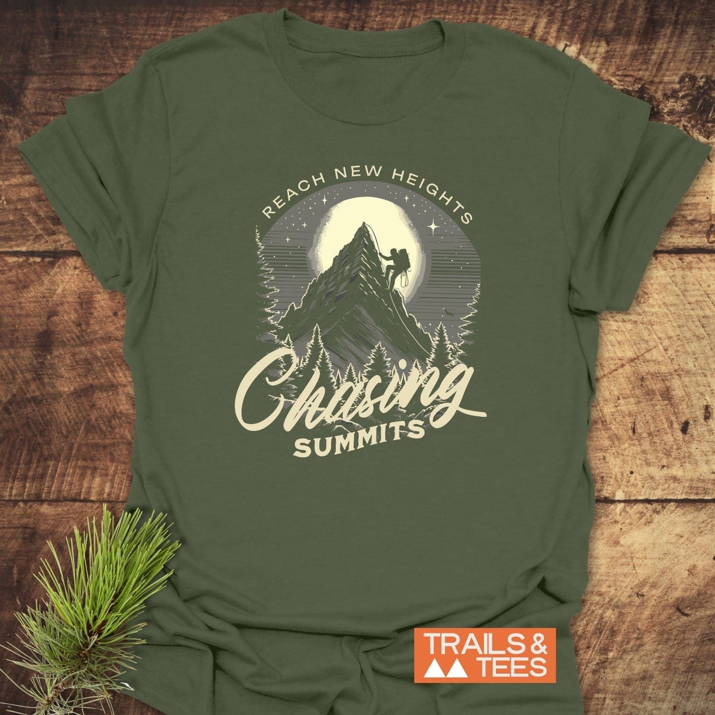 Chasing Summits T-Shirt featuring a climber graphic on an olive-green shirt, highlighting outdoor adventure themes. Displayed on rustic wood with a pine sprig accent.