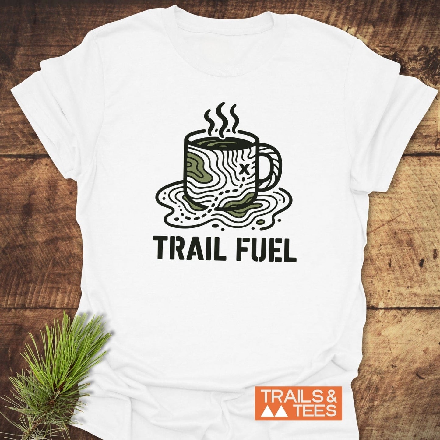 Trail Fuel T-Shirt displaying a coffee mug over trail map design, TRAIL FUEL text, on a wood-grain background with a pine sprig nearby.