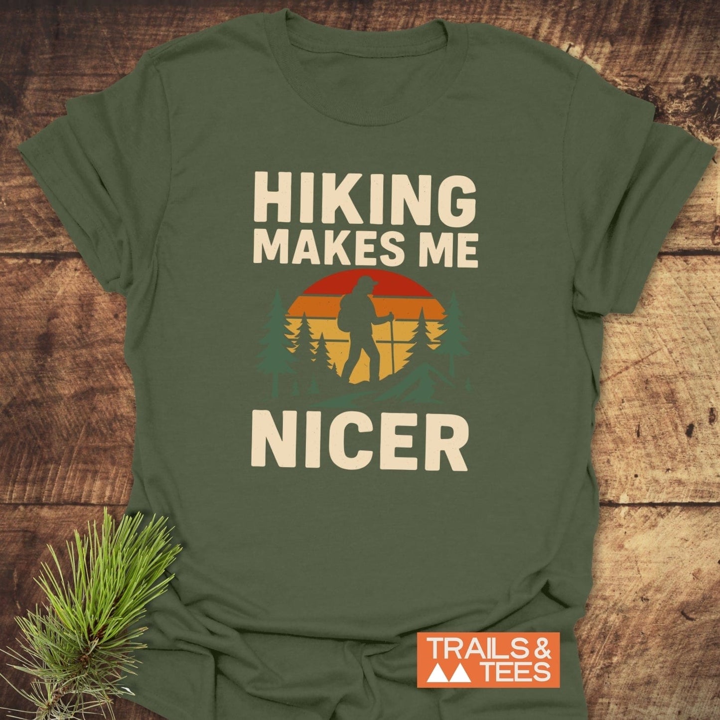 Hiking Makes Me Nicer T-Shirt featuring a retro hiker graphic, crafted from soft ring-spun cotton, laid on wood with a pine sprig beside.
