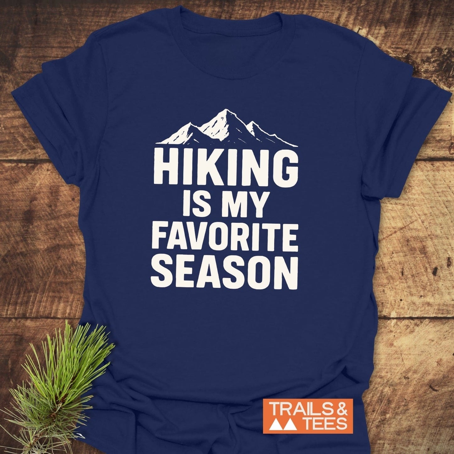 Navy-blue Hiking Is My Favorite Season T-shirt with mountain design, laid flat on wood, featuring Trails & Tees logo and pine needles for outdoor touch.