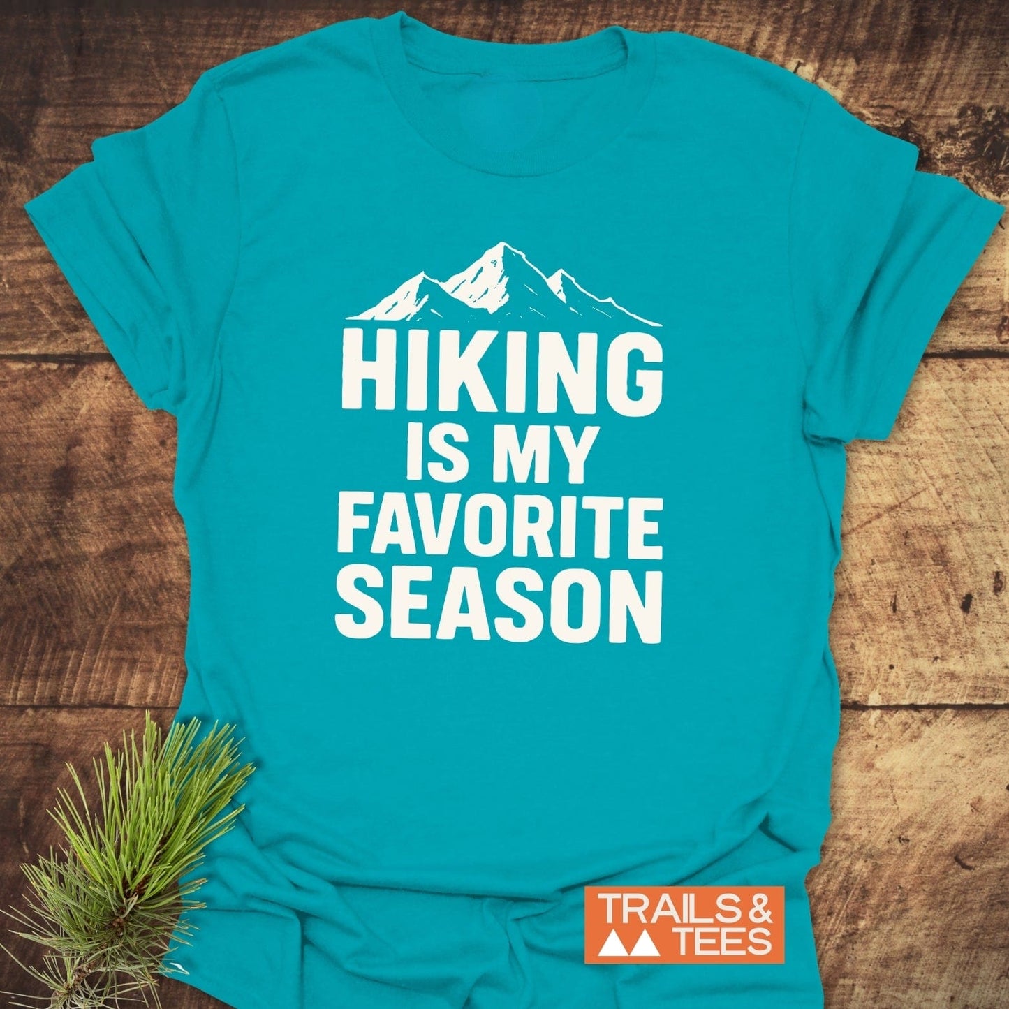 Hiking Is My Favorite Season T-Shirt with mountain silhouette, orange TRAILS & TEES label, and pine needles, highlighting its outdoor adventure design and comfort.