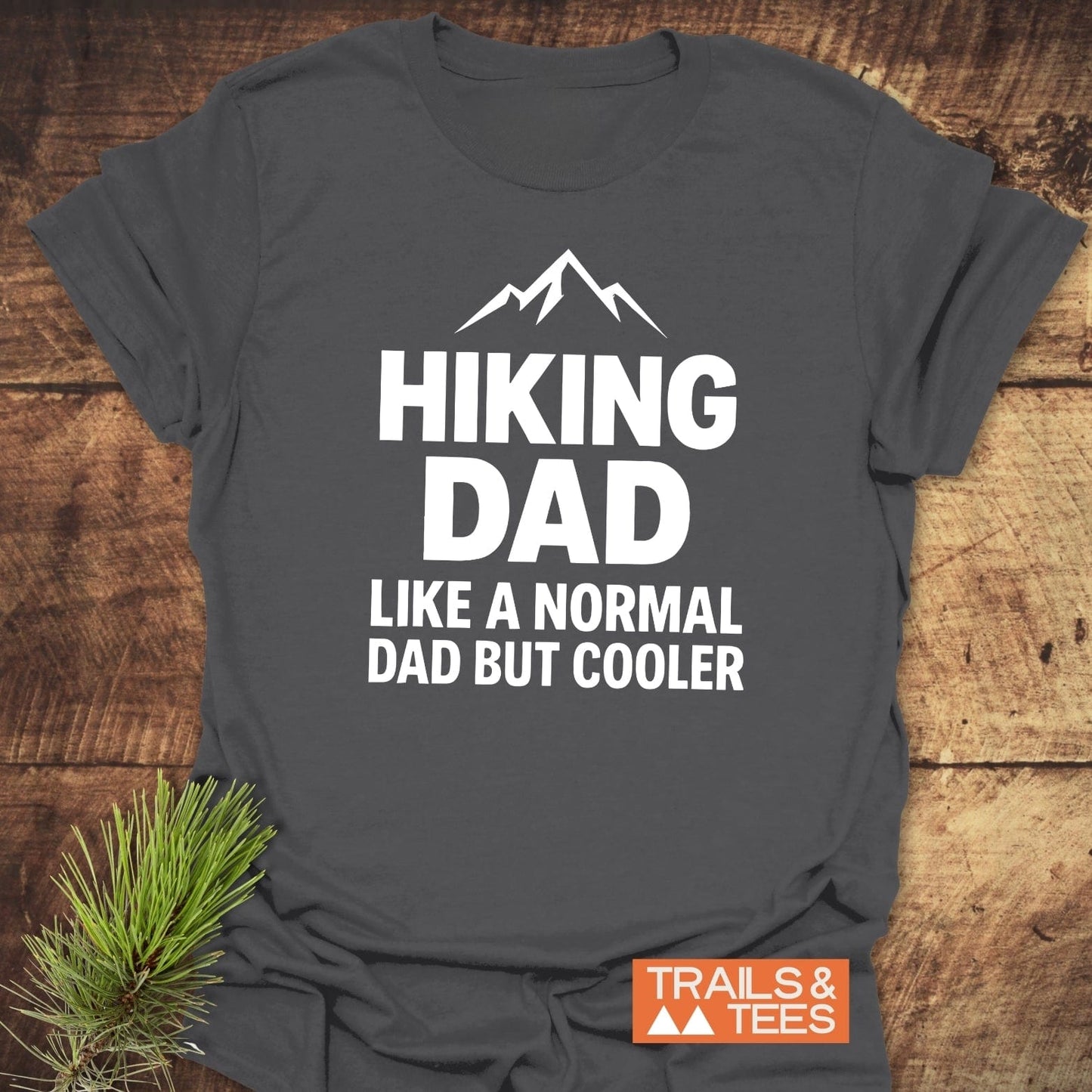 Hiking Dad Funny T-Shirt in dark gray with bold white print and mountain silhouette, laid flat with a pine sprig beside it, showcasing rustic outdoor style.