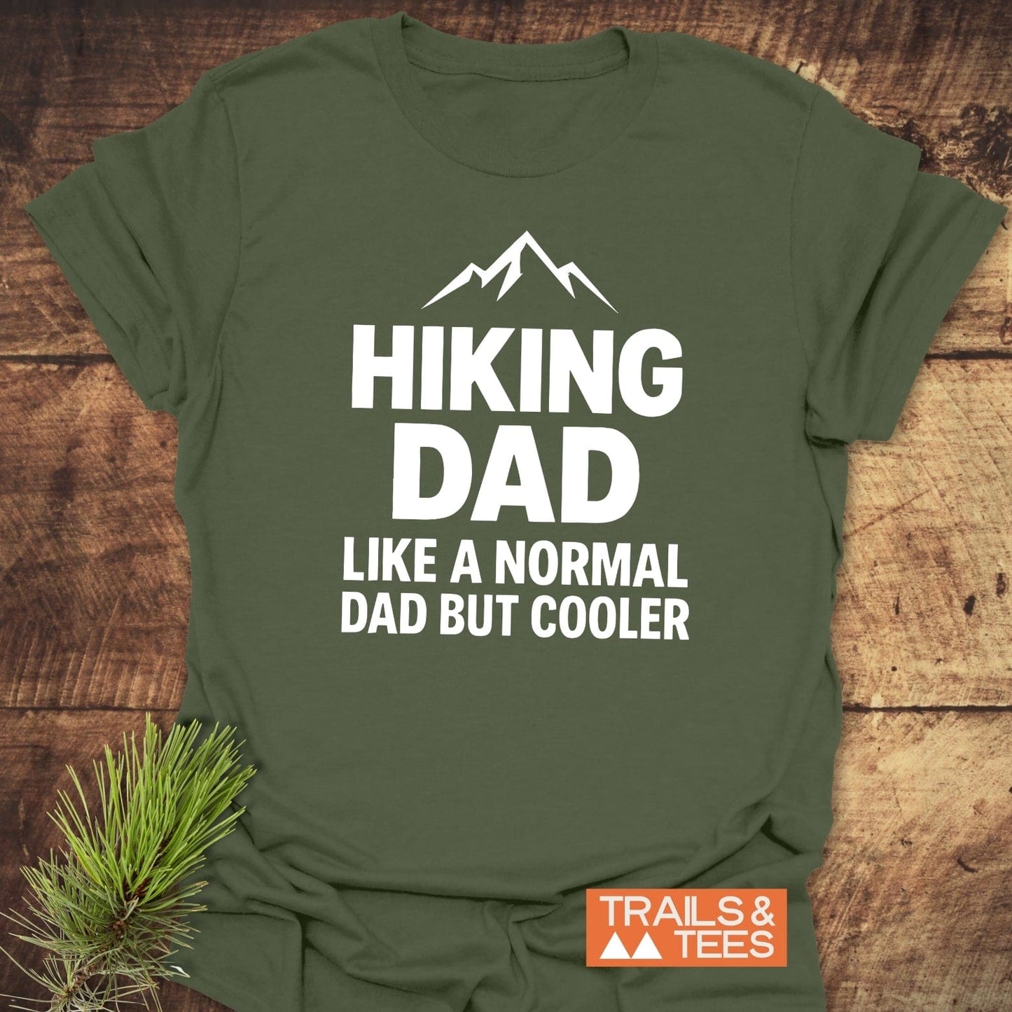 Olive-green Hiking Dad T-shirt with mountain graphic, bold white text, and Trails & Tees logo, showcasing outdoor comfort and style, laid on wood.