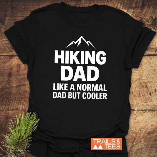 Black Hiking Dad T-shirt with mountain graphic, featuring Like a Normal Dad but Cooler text; soft, pre-shrunk cotton, durable print, laid on wood surface.