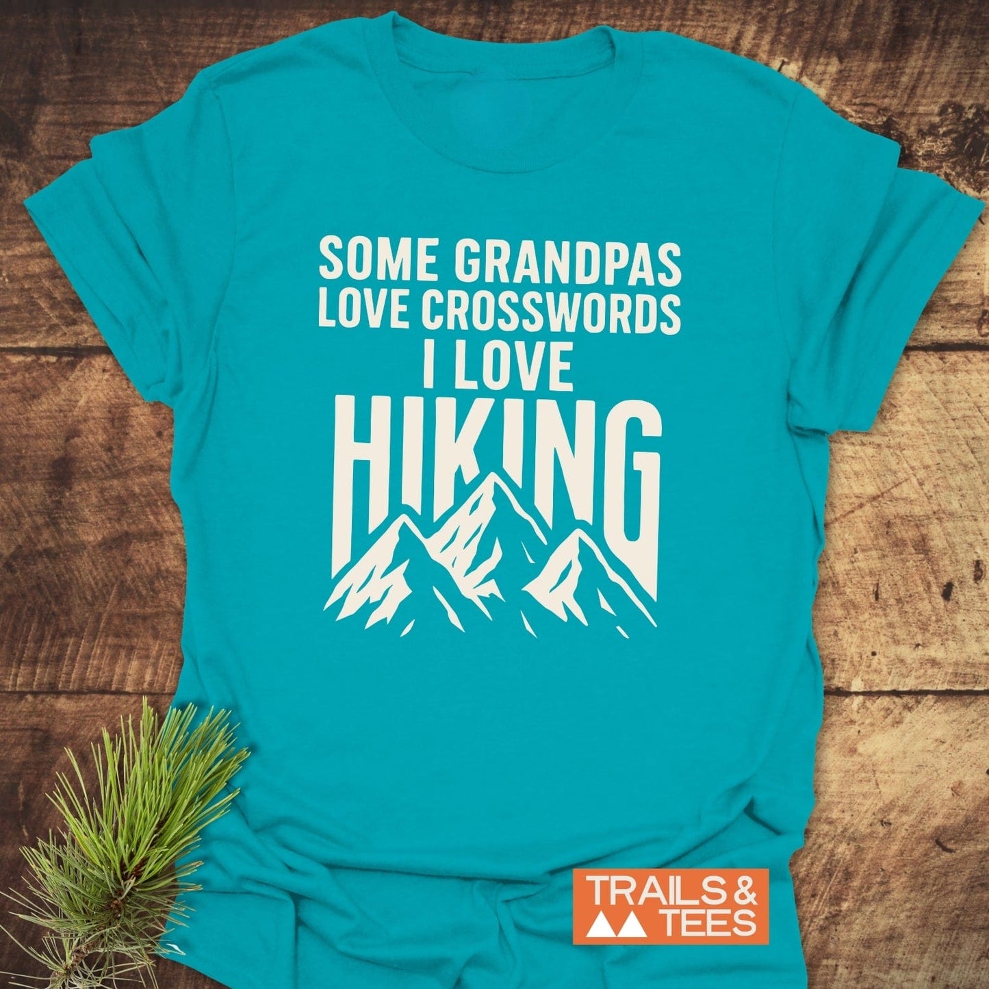Grandpa Loves Hiking Funny T-Shirt, featuring white mountain peaks and text, laid flat with orange “TRAILS & TEES” logo, pine sprig accent on wooden surface.