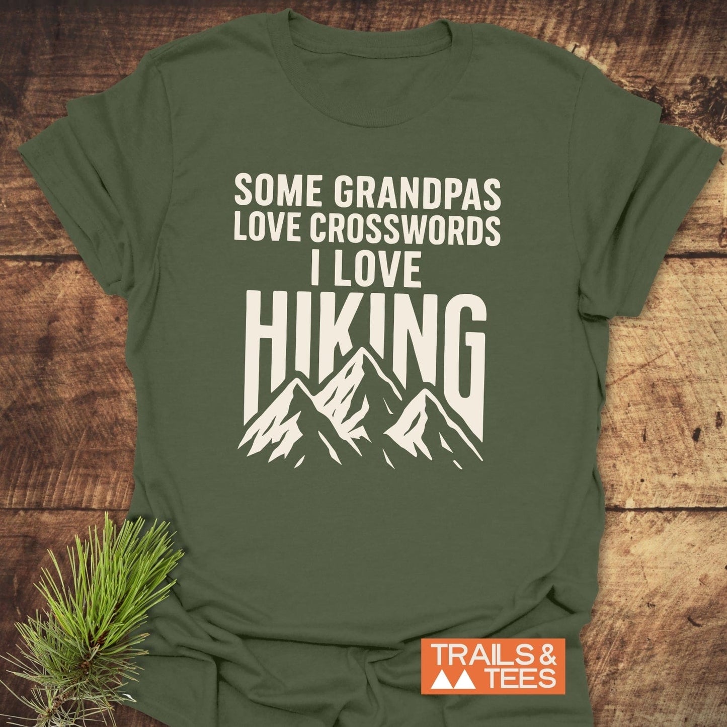 Grandpa Loves Hiking Funny T-Shirt featuring a mountain graphic and slogan, styled with a pine sprig on wood surface, showcasing outdoor-themed design and comfort.