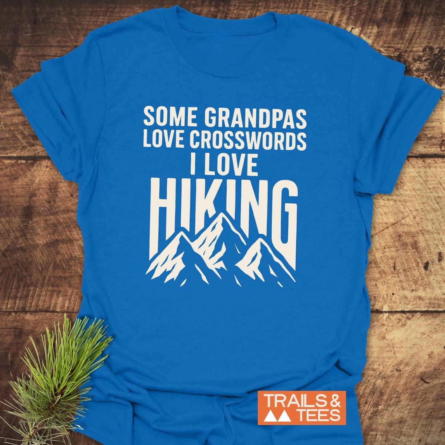 Grandpa Loves Hiking Funny T-Shirt with bold slogan and mountain graphic, laid on wood surface with pine sprig, showcasing outdoor adventure spirit.