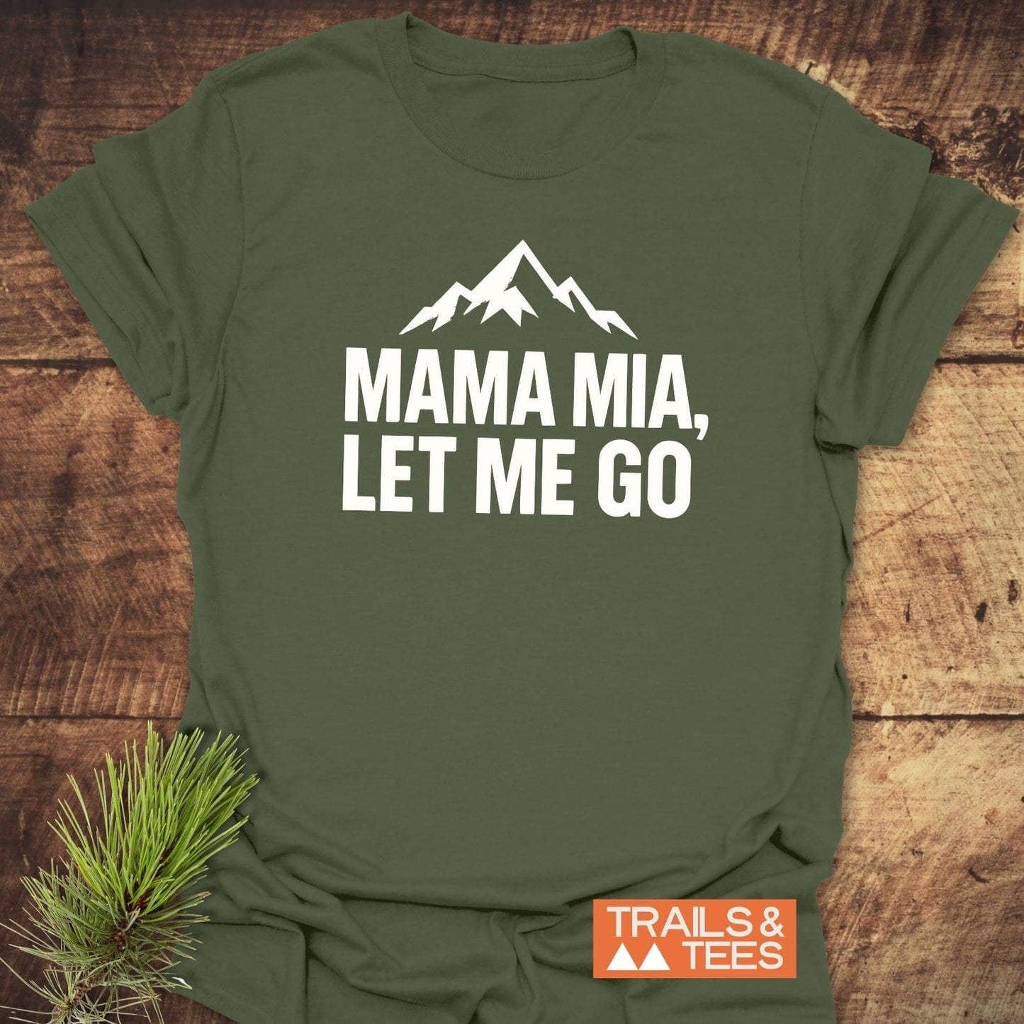Let Me Go Funny T-Shirt featuring a mountain silhouette and phrase, laid flat with a pine sprig and Trails & Tees logo visible.