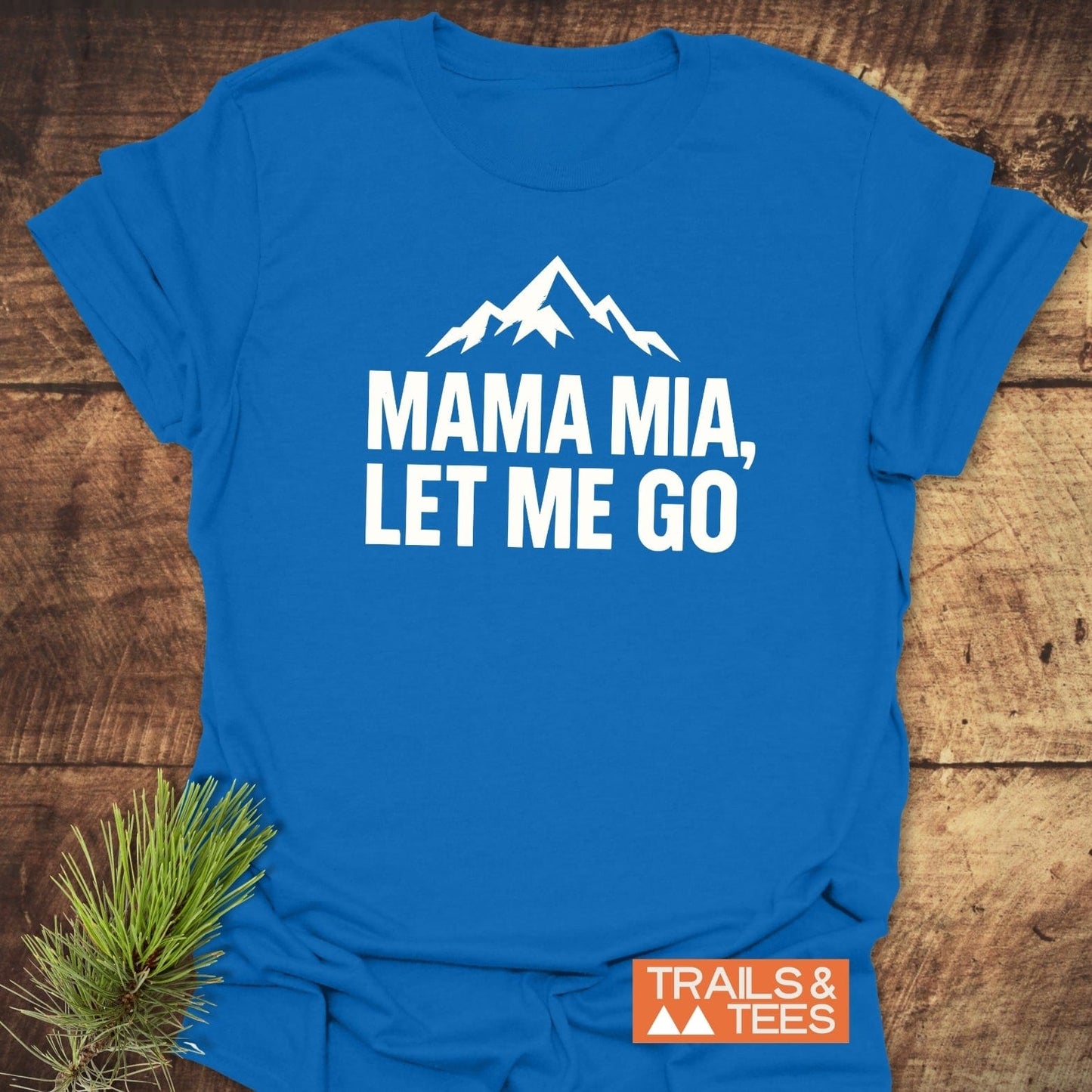 Let Me Go Funny T-Shirt with mountain graphic, MAMA MIA, LET ME GO text, orange Trails & Tees logo, displayed on wood with pine twig accent.