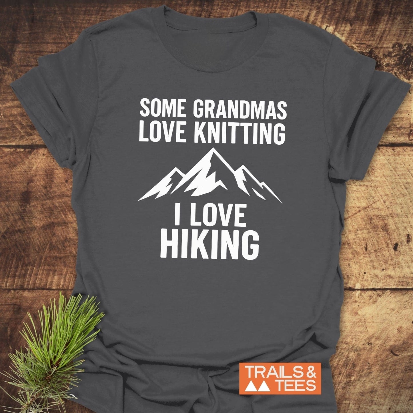 Dark-gray Hiking Grandma Funny T-Shirt with white mountain graphic and slogan, Some Grandmas Love Knitting, I Love Hiking, on a wooden surface.