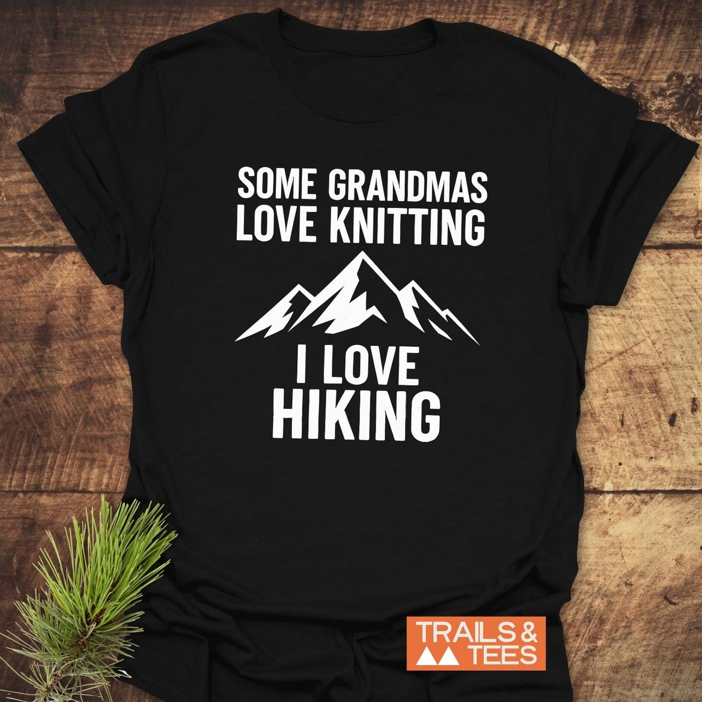 Black T-shirt with a white mountain graphic and Some Grandmas Love Knitting, I Love Hiking slogan. Trails & Tees logo. Perfect for outdoor enthusiasts.